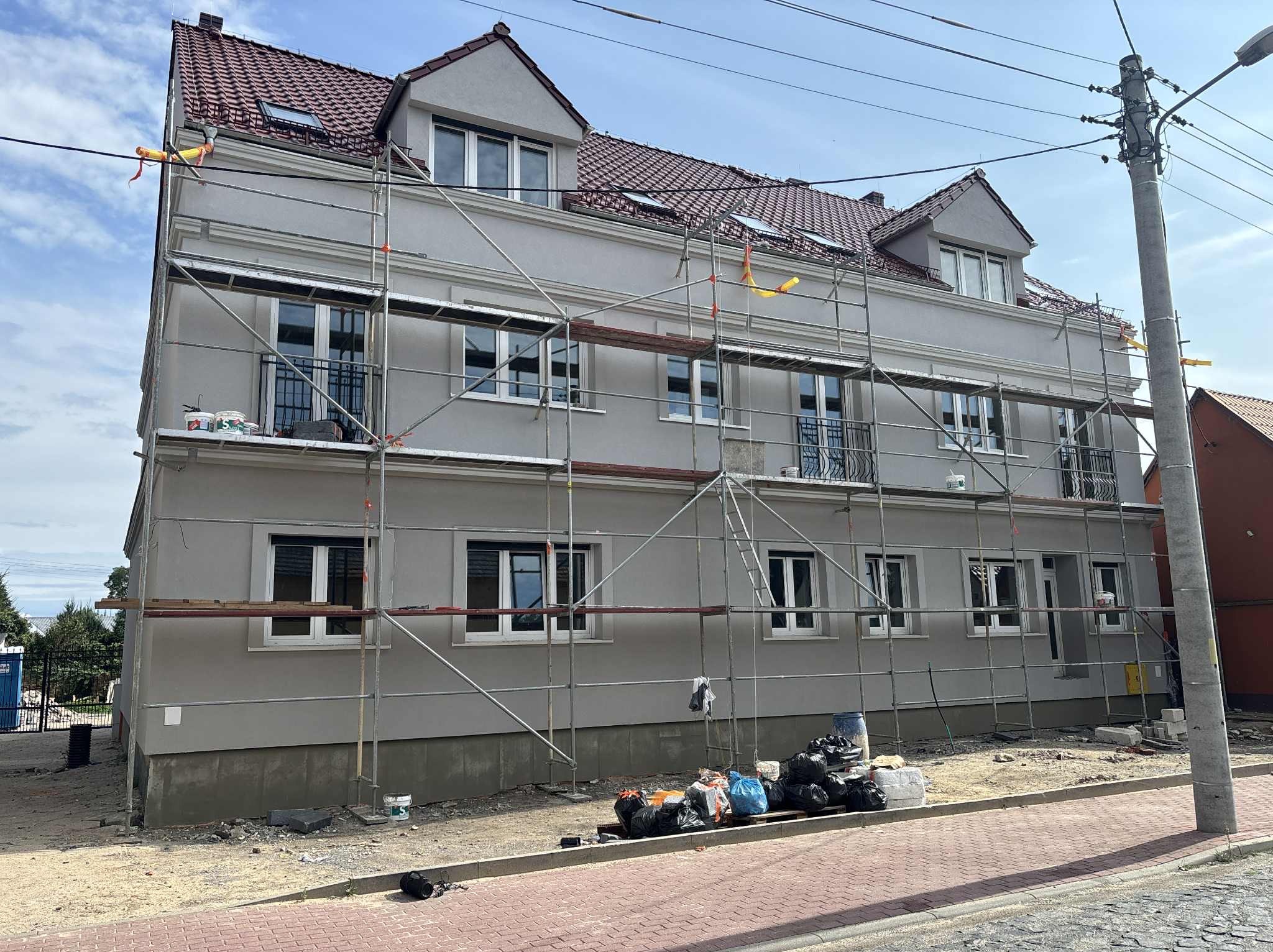 Budynek wielorodzinny w trakcie prac elewacyjnych, widoczne rusztowania i worki z odpadami budowlanymi. Świeżo położona szara elewacja z białymi oknami.
