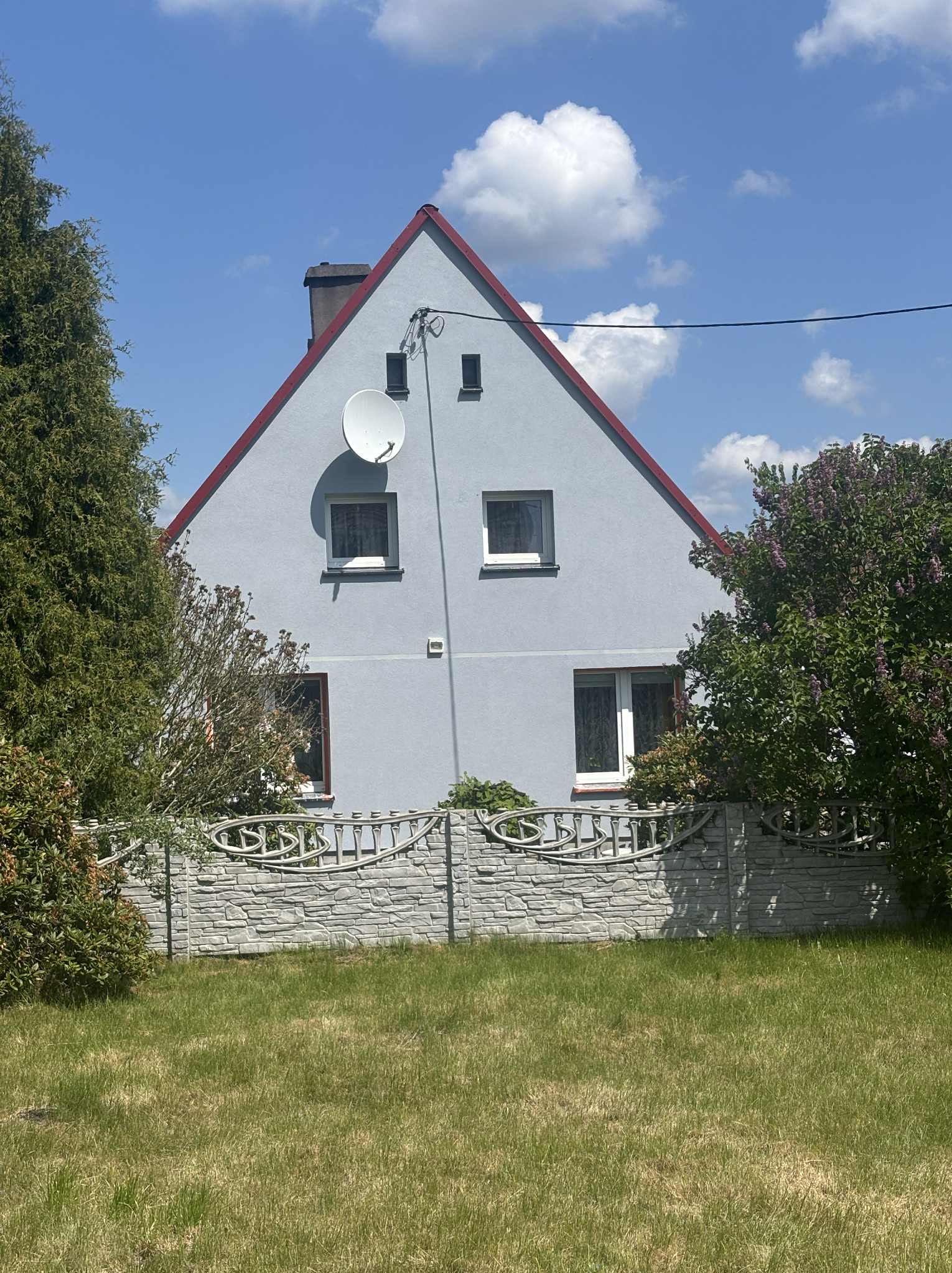 Szary dom jednorodzinny z czerwoną obwódką dachu, widoczny za ozdobnym, betonowym ogrodzeniem. Błękitne niebo z chmurami w tle.