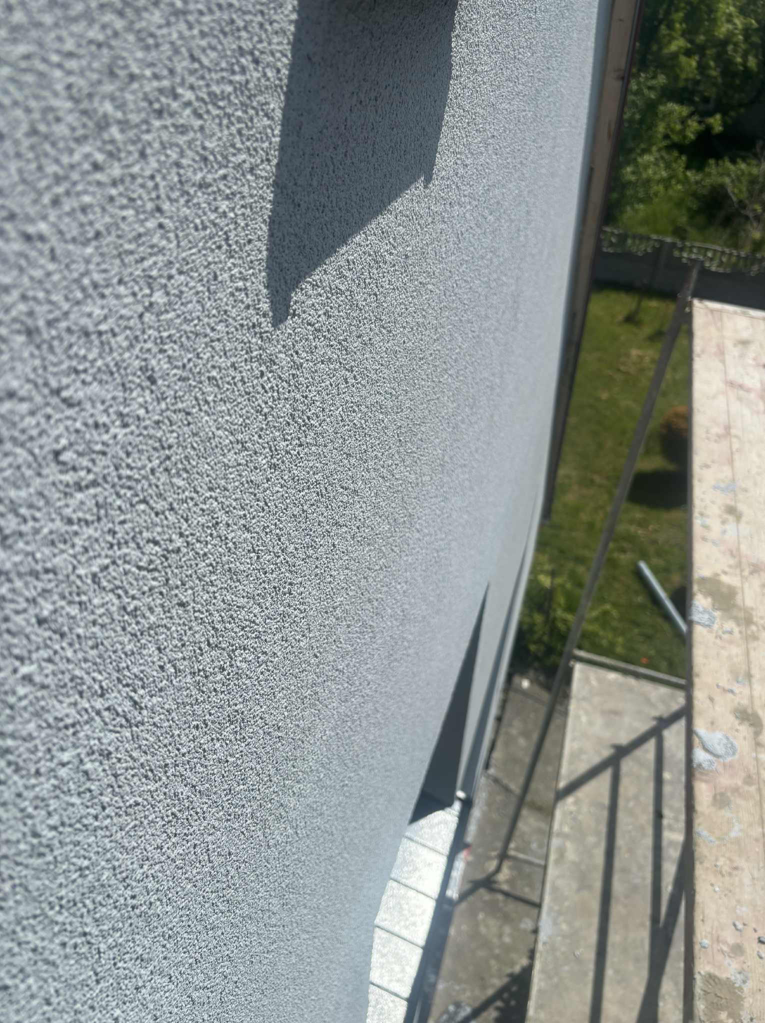 Szara, ziarnista elewacja budynku z bliska, widziana z rusztowania. Świeżo nałożony tynk strukturalny w neutralnym odcieniu szarości.