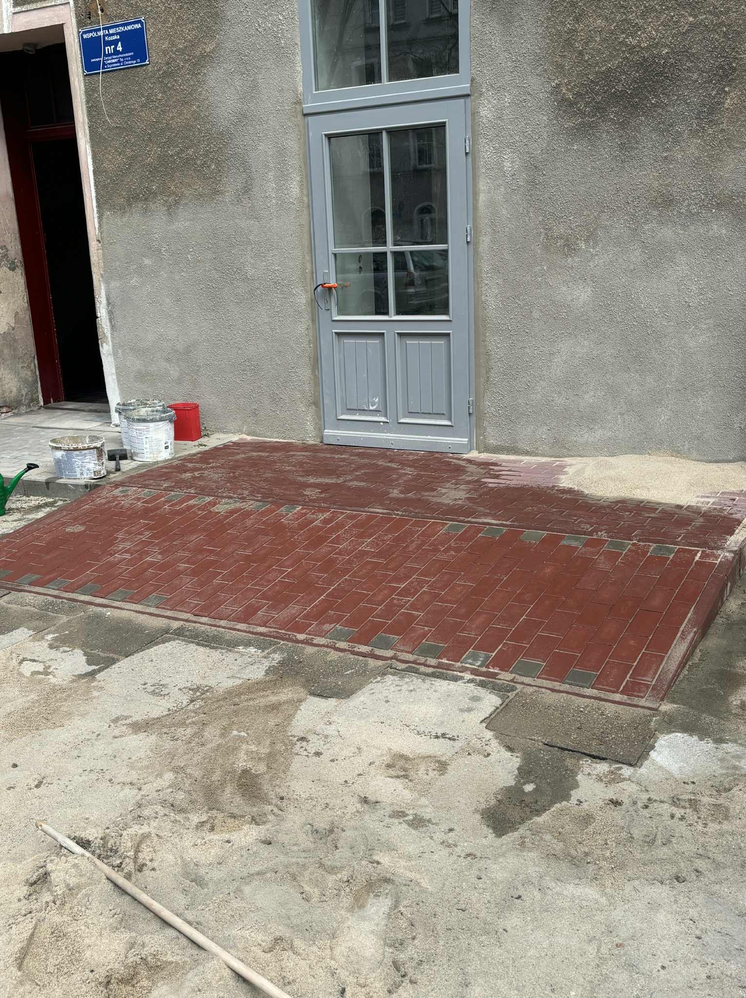 Świeżo ułożona, czerwona kostka brukowa przed wejściem do budynku. Widoczne ślady piasku i narzędzia. Szara elewacja i drzwi wejściowe.
