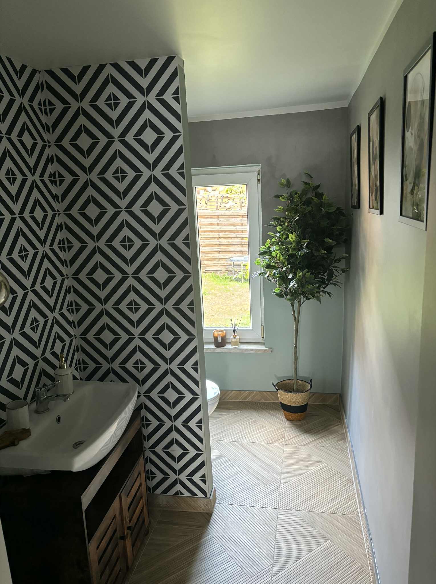 Wzorzysta tapeta w romby w łazience w Iłowej. Umywalka z szafką, okno z widokiem na zewnątrz, podłoga w jasne pasy. Dekoracyjne drzewko w rogu.