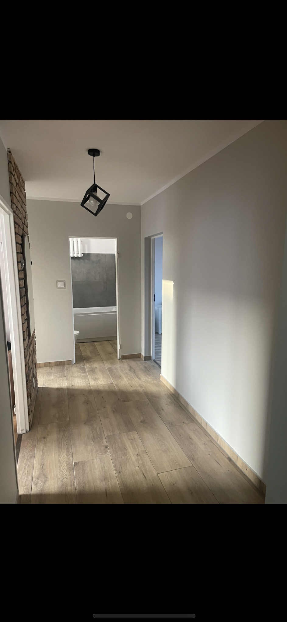 Jasne panele podłogowe w korytarzu z szarą ścianą i ozdobną cegłą. W tle widoczna łazienka z wanną. Geometryczna lampa sufitowa dodaje nowoczesnego akcentu.