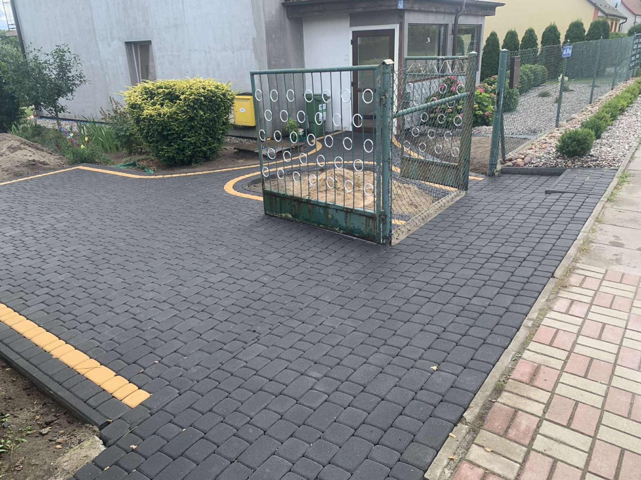 Ułożona szara kostka brukowa z żółtymi akcentami przy posesji z zielonym metalowym ogrodzeniem i bramą. Widoczny fragment budynku i roślinność.