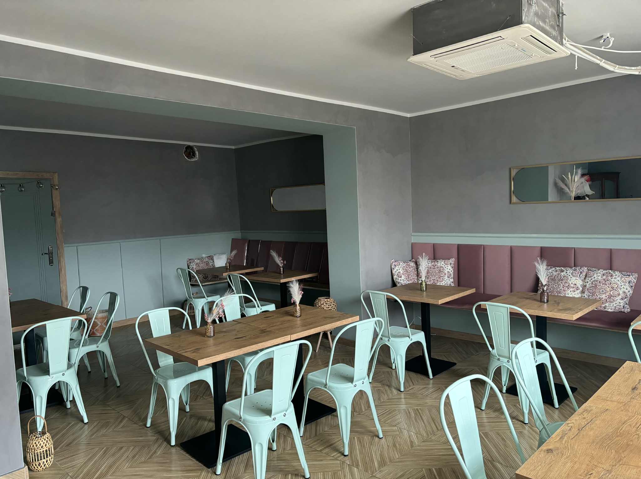 Wnętrze lokalu po malowaniu: pastelowe ściany, drewniane stoły i miętowe krzesła. Dekoracyjne detale, w tym lustra i tekstylia, dopełniają nowoczesny, minimalistyczny styl.