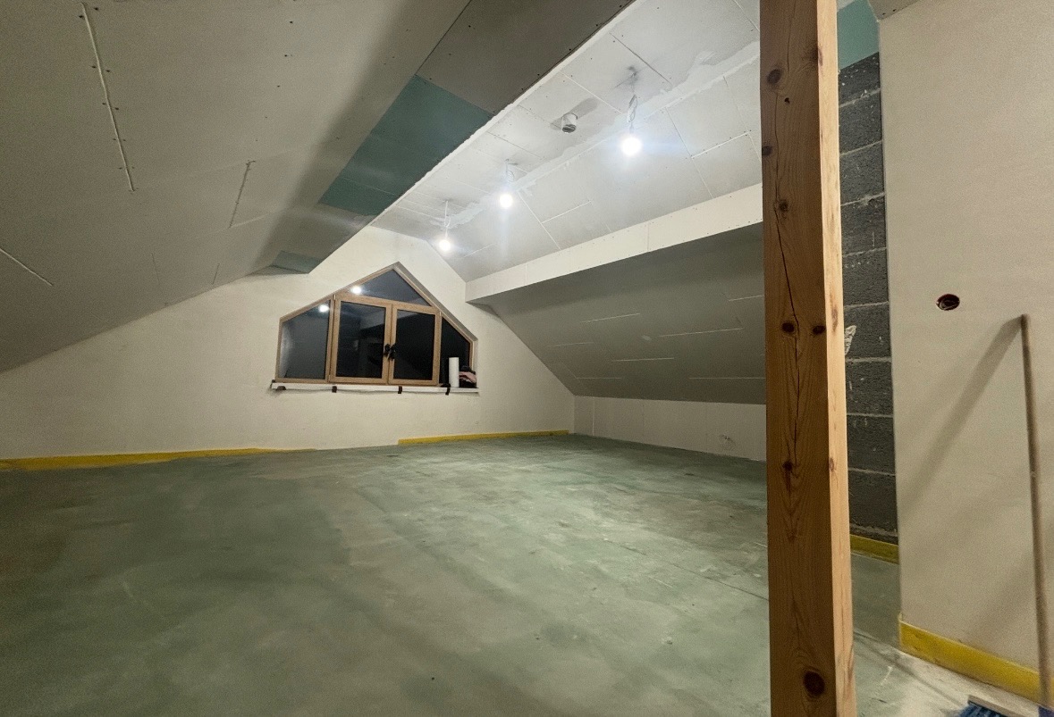 Poddasze ze skośnym oknem, przygotowane do prac wykończeniowych. Widoczne płyty gipsowe na ścianach i suficie, drewniany słup konstrukcyjny. Podłoga zabezpieczona folią.