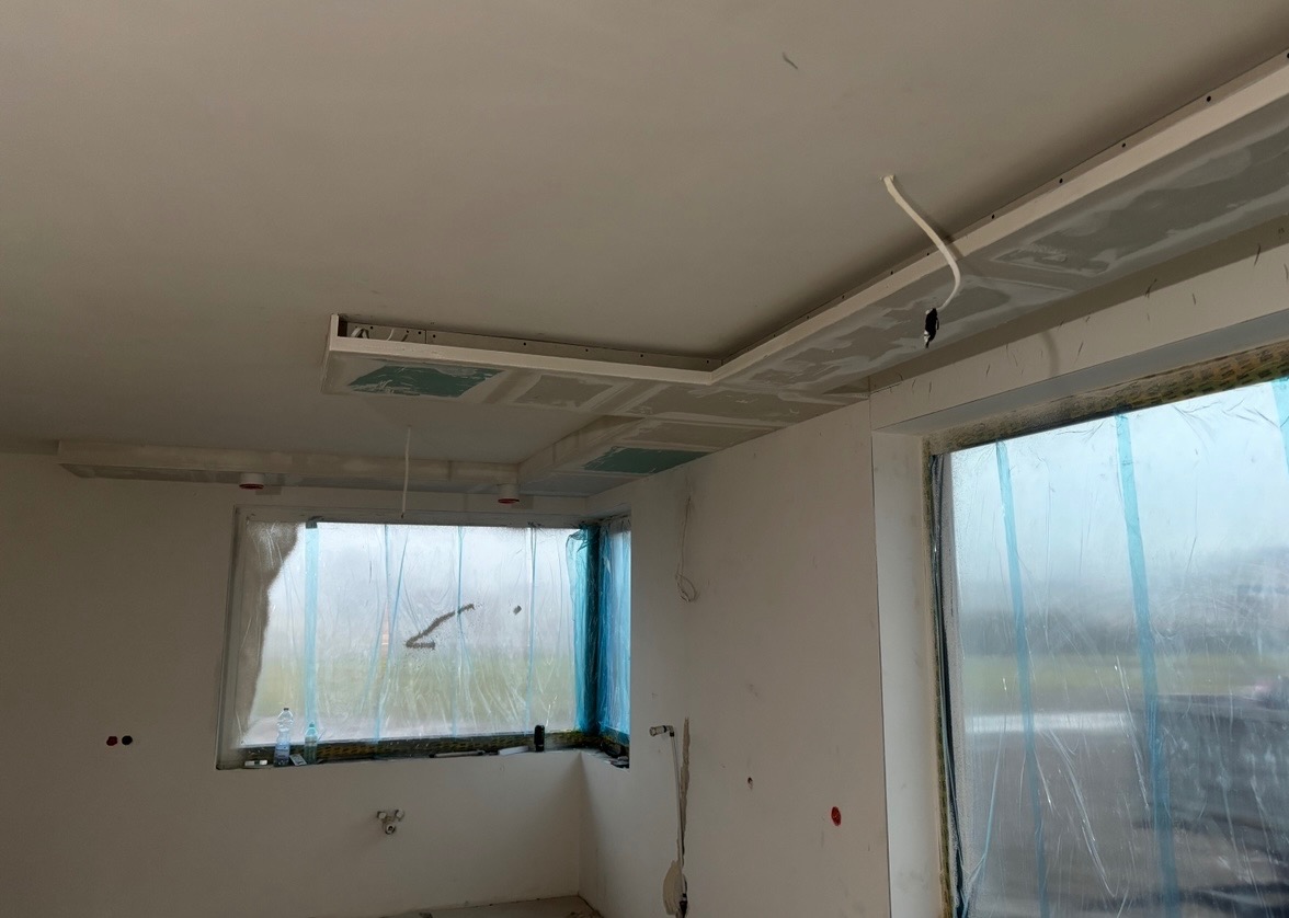 Wnętrze w trakcie prac wykończeniowych: zabudowa sufitu z płyt gipsowo-kartonowych, okna zabezpieczone folią. Widoczne przewody elektryczne i elementy instalacji.