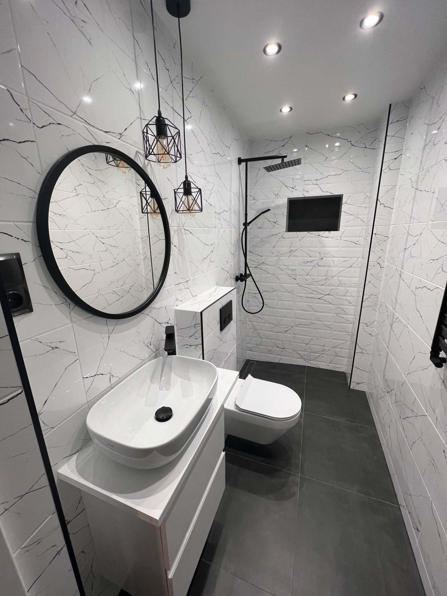 Nowoczesna łazienka w Sandomierzu po remoncie, z białymi płytkami imitującymi marmur, czarną armaturą, okrągłym lustrem i wiszącymi lampami w stylu industrialnym.