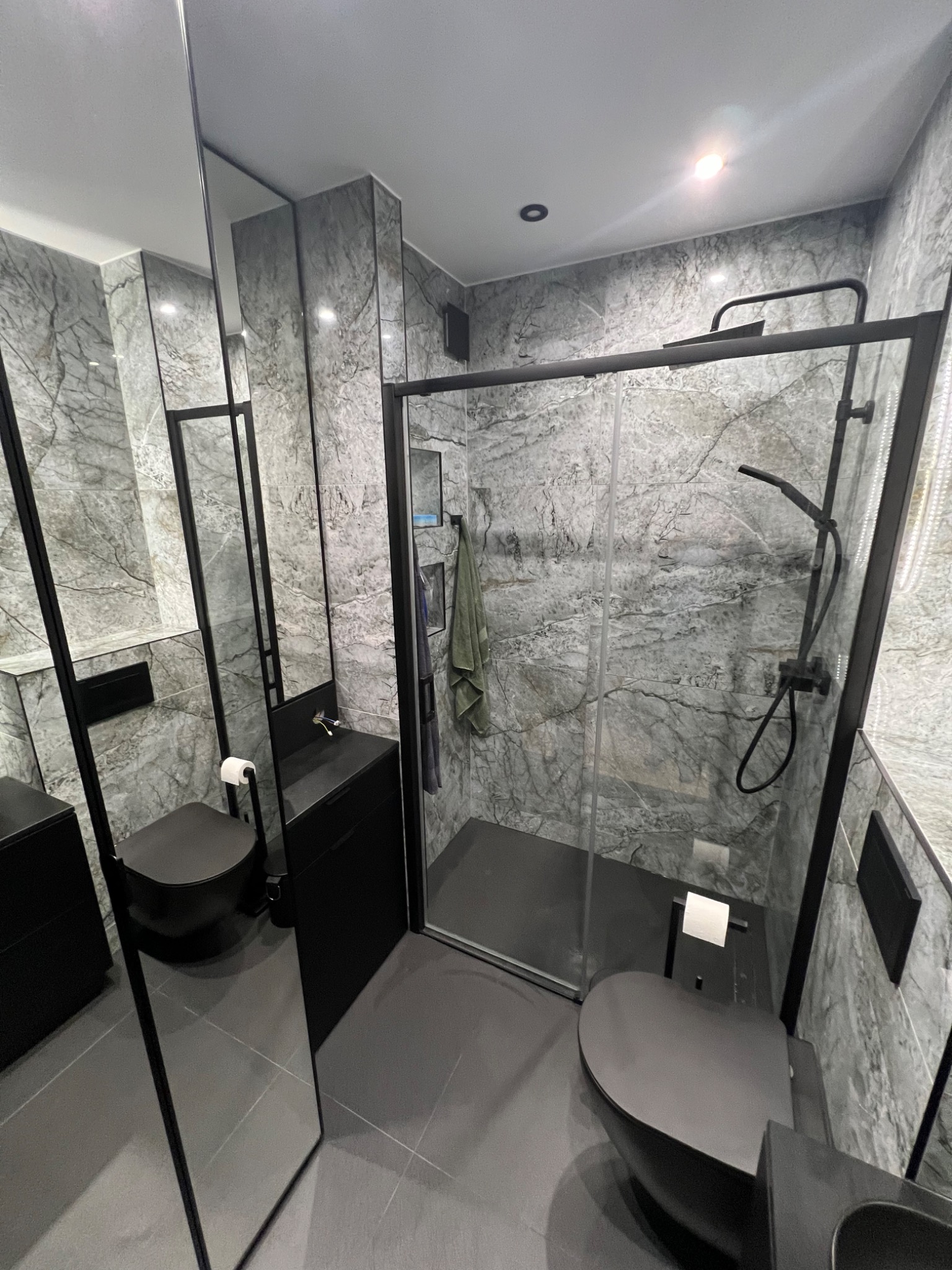Nowoczesna łazienka z szarymi marmurkowymi płytkami, czarną armaturą i kabiną prysznicową ze szklanymi drzwiami, widok na toaletę i umywalkę, minimalistyczny design z wiszącą toaletą i ręcznikiem...