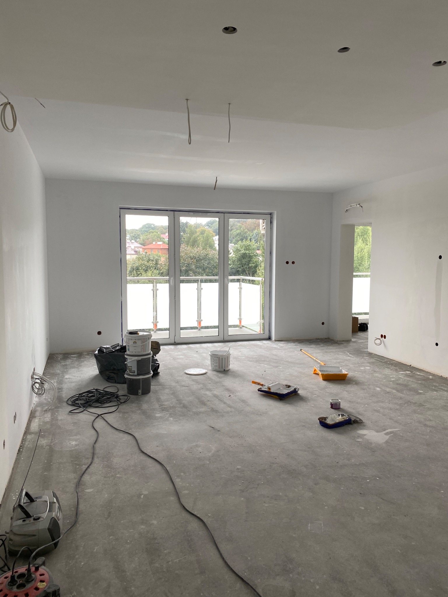 Wnętrze w trakcie odświeżania: świeżo pomalowane na biało ściany i sufit, widoczne puszki z farbą, wałki malarskie, kable zwisające z sufitu, surowa podłoga przed położeniem wykończenia.
