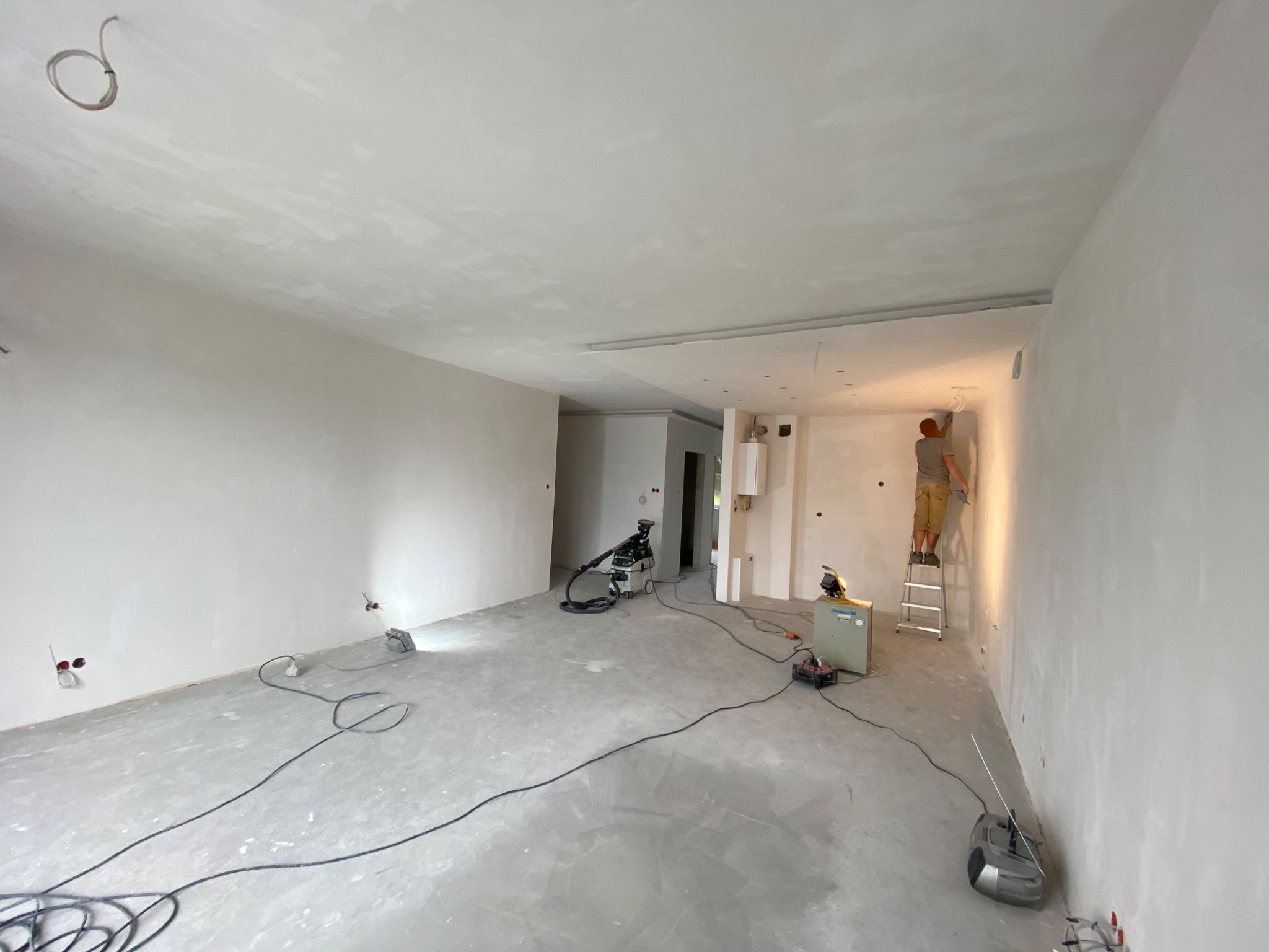 Przestronne wnętrze w trakcie prac wykończeniowych: ściany po szpachlowaniu, widoczne kable elektryczne, drabina z pracownikiem przy suficie, sprzęt budowlany na podłodze, surowy betonowy podkład.