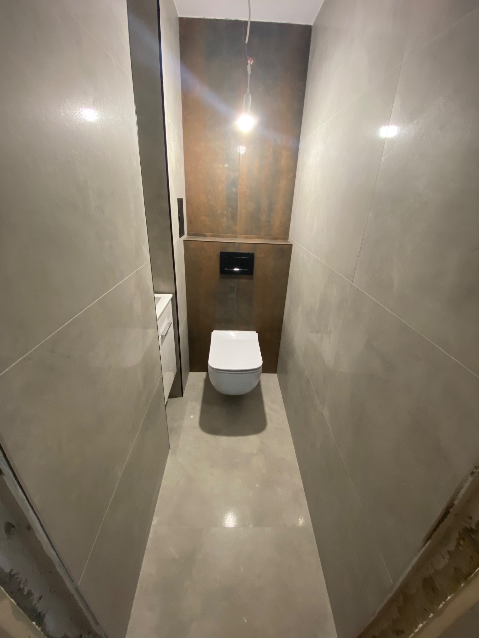 Wąska toaleta w szaro-brązowej kolorystyce, z wiszącą muszlą klozetową i ukrytym spłukiwaniem, oświetlona pojedynczą żarówką.