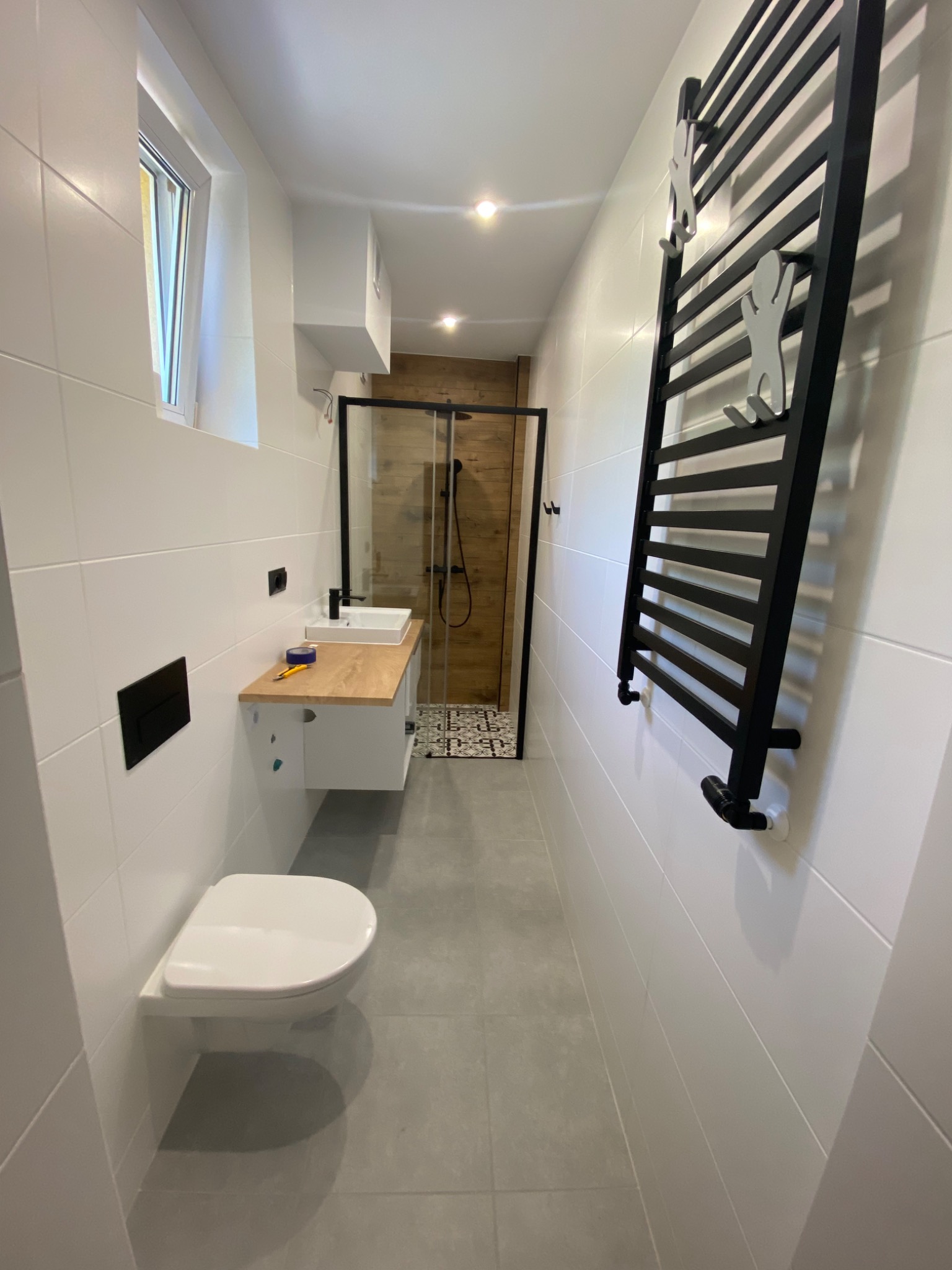 Wąska łazienka z białymi płytkami, wiszącą toaletą, umywalką na drewnianym blacie i czarną kabiną prysznicową z drewnianą ścianą w tle, grzejnik drabinkowy z figurkami wspinaczy.