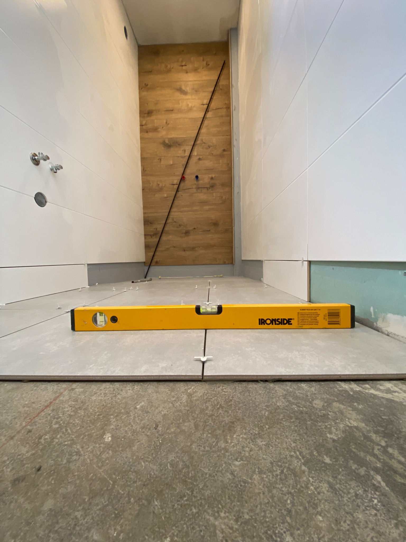 Poziomica Ironside na szarych płytkach podłogowych w trakcie układania, białe płytki ścienne częściowo ułożone, drewnopodobna ściana w głębi, widoczne krzyżyki dystansowe.