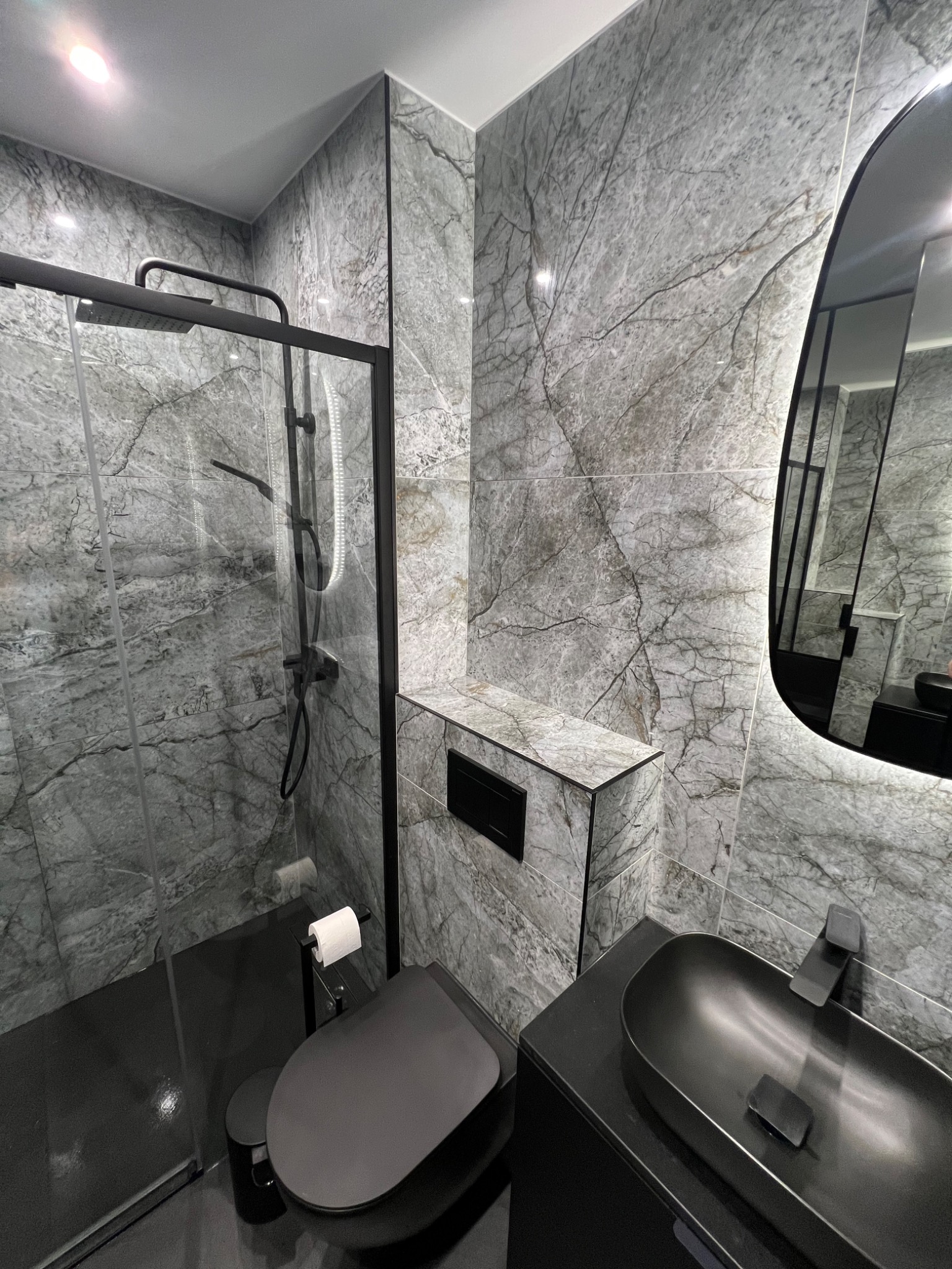 Nowoczesna łazienka w odcieniach szarości z czarnymi akcentami: kabina prysznicowa typu walk-in, wisząca toaleta, umywalka nablatowa i podświetlane lustro owalne.