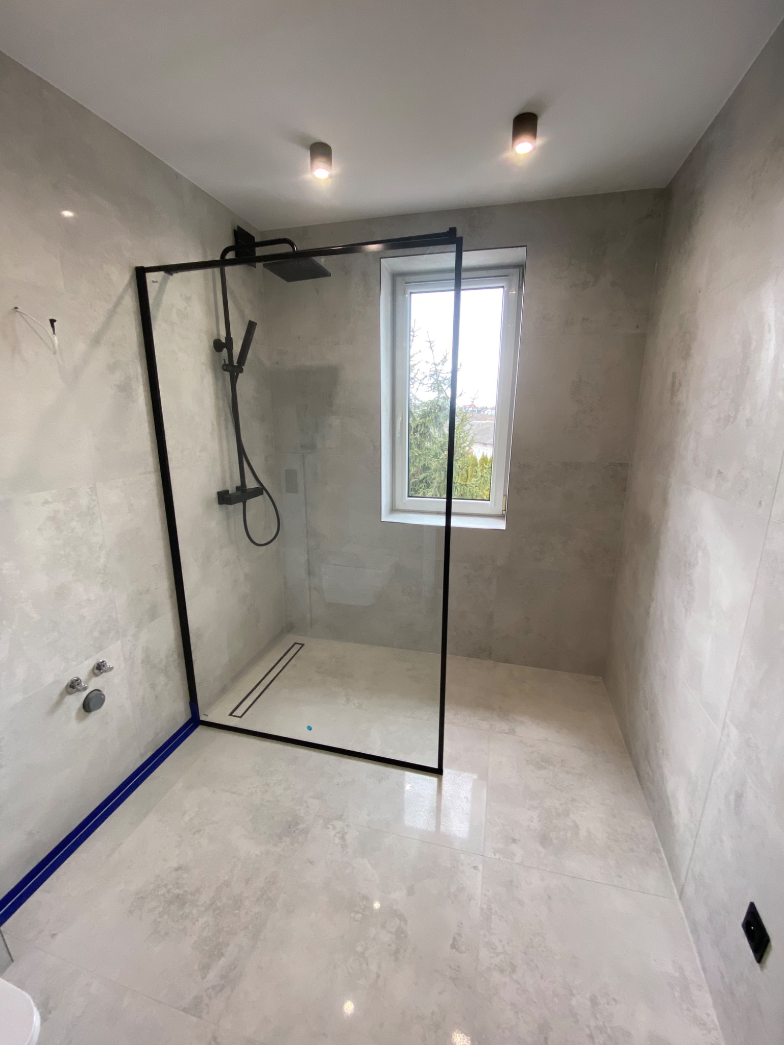 Nowoczesna łazienka z kabiną prysznicową typu walk-in z czarną ramą, szarymi płytkami imitującymi beton i oknem z widokiem na zieleń.