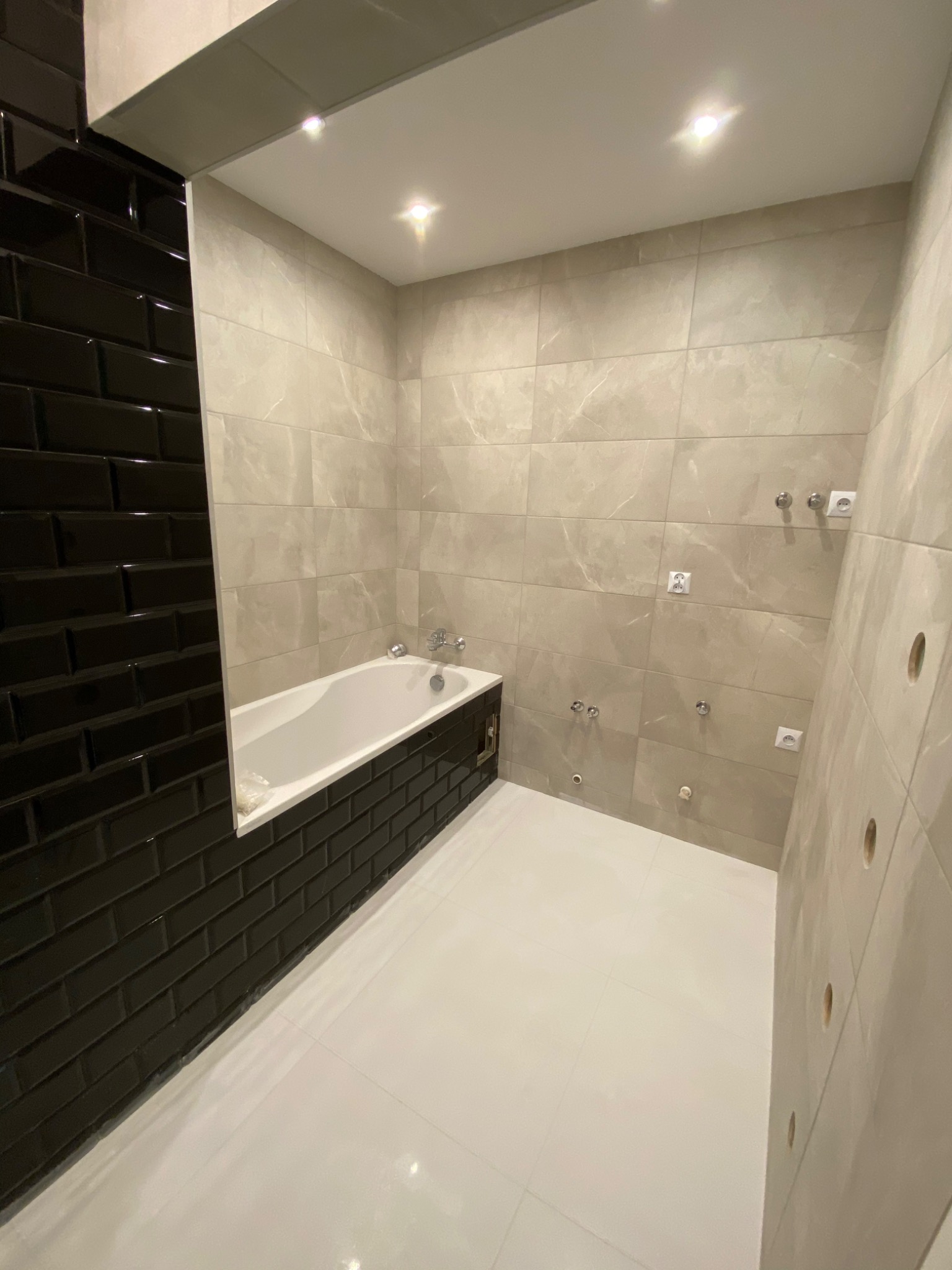 Wykończona łazienka z wanną obudowaną czarnymi, błyszczącymi płytkami typu metro, jasne płytki na ścianach z widocznymi puszkami elektrycznymi i wyprowadzeniami hydraulicznymi, biała podłoga...