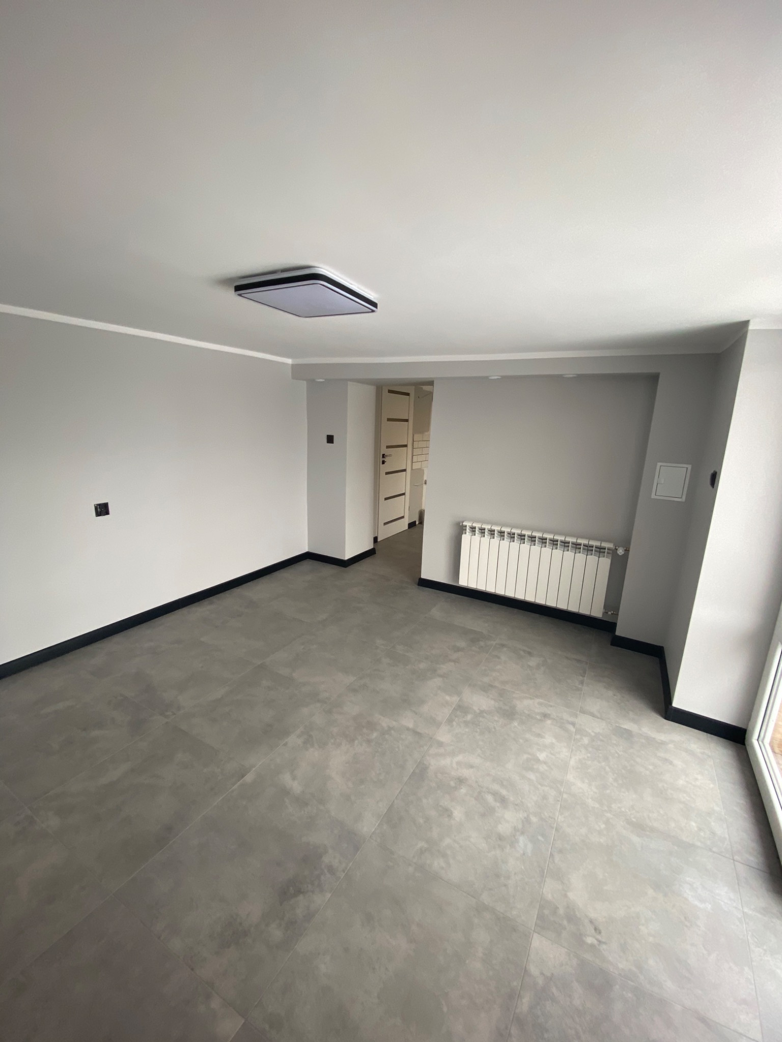 Nowoczesne, puste wnętrze z szarymi ścianami, płytkami podłogowymi imitującymi beton, białym grzejnikiem i minimalistycznym oświetleniem sufitowym.