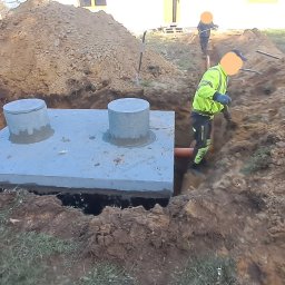 DSF Wyburzenia-Rozbiórki Dominik Filipek - Montaż betonowego zbiornika z dwoma włazami w wykopie, widoczny pracownik w odblaskowej kamizelce i pomarańczowa rura odpływowa, w tle koparka i sterta ziemi.