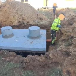 Montaż betonowego zbiornika z dwoma włazami w wykopie, widoczny pracownik w odblaskowej kamizelce i pomarańczowa rura odpływowa, w tle koparka i sterta ziemi.