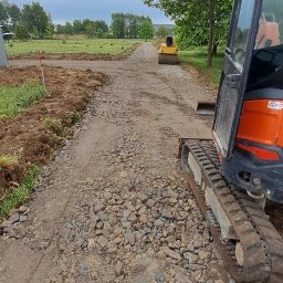 DSF Wyburzenia-Rozbiórki Dominik Filipek - Utwardzanie drogi gruntowej z użyciem walca i minikoparki. Widoczne świeżo rozplantowane kamienie i ziemia. Prace w toku, sprzęt budowlany w użyciu.