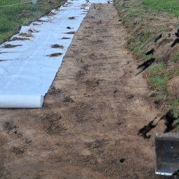 DSF Wyburzenia-Rozbiórki Dominik Filipek - Przygotowanie podłoża pod drogę: rozłożona biała geowłóknina na wyrównanym terenie, widok z kabiny maszyny budowlanej, w tle zabudowania.