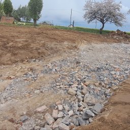 DSF Wyburzenia-Rozbiórki Dominik Filipek - Przygotowanie terenu pod budowę drogi: wyrównany teren z gruzem i kamieniami, w tle drzewa owocowe i pole rzepaku. Widoczne ślady opon traktora.