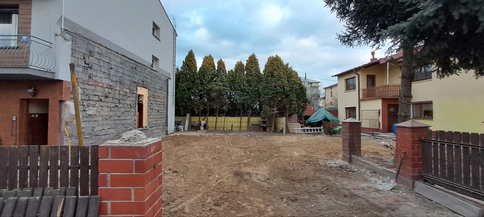 Przygotowany teren pod budowę z widocznymi śladami opon, częściowo rozebrana ściana budynku z cegły i elementy nowego ogrodzenia z cegły klinkierowej.