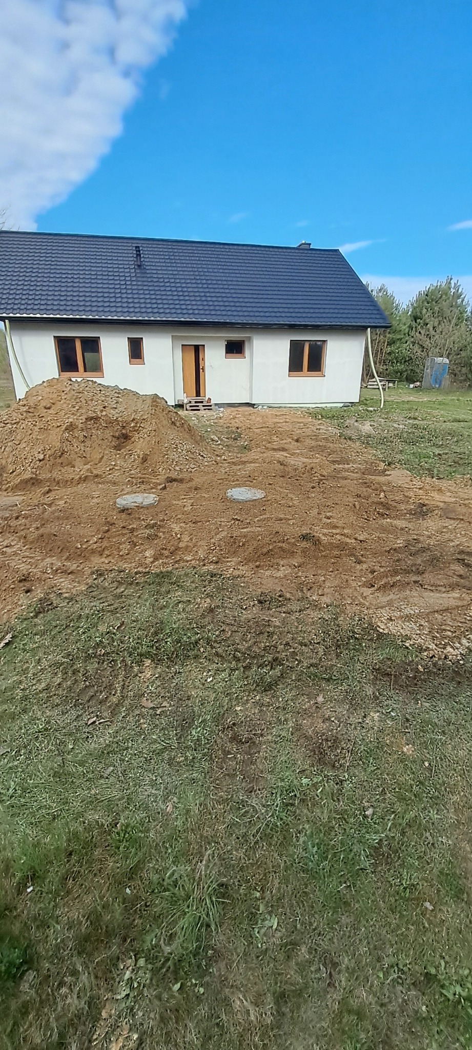 Nierówny teren przed nowo wybudowanym domem z widocznymi studzienkami kanalizacyjnymi i stertą ziemi.