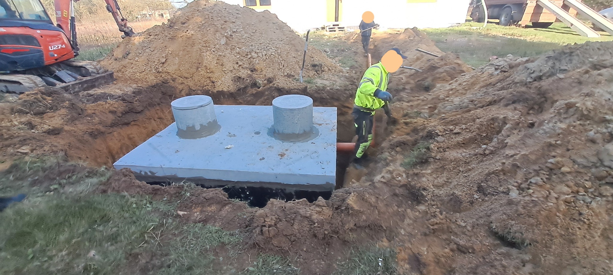 Montaż betonowego zbiornika z dwoma włazami w wykopie, widoczny pracownik w odblaskowej kamizelce i pomarańczowa rura odpływowa, w tle koparka i sterta ziemi.