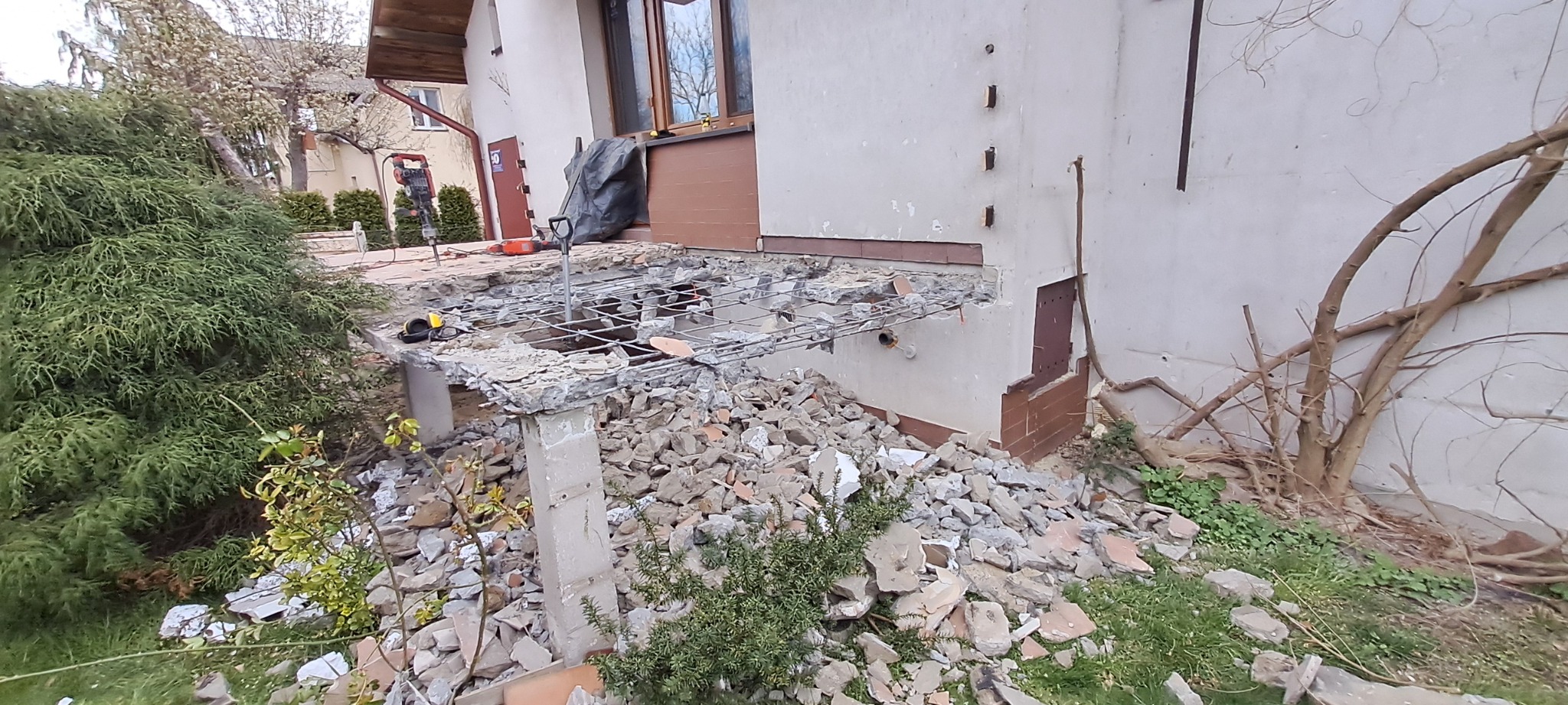 Demontaż betonowej konstrukcji tarasu przy budynku jednorodzinnym, widoczne zbrojenie, gruz i narzędzia budowlane.