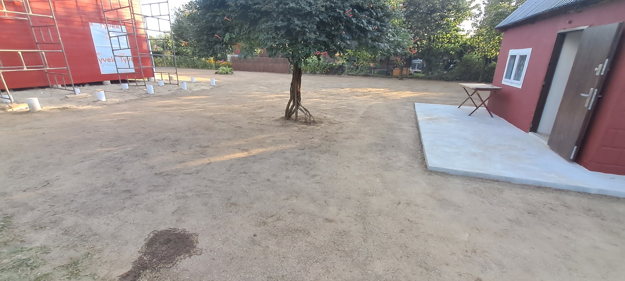 Teren po niwelacji z widocznym budynkiem w trakcie budowy i drzewem. Przygotowany grunt pod dalsze prace budowlane.