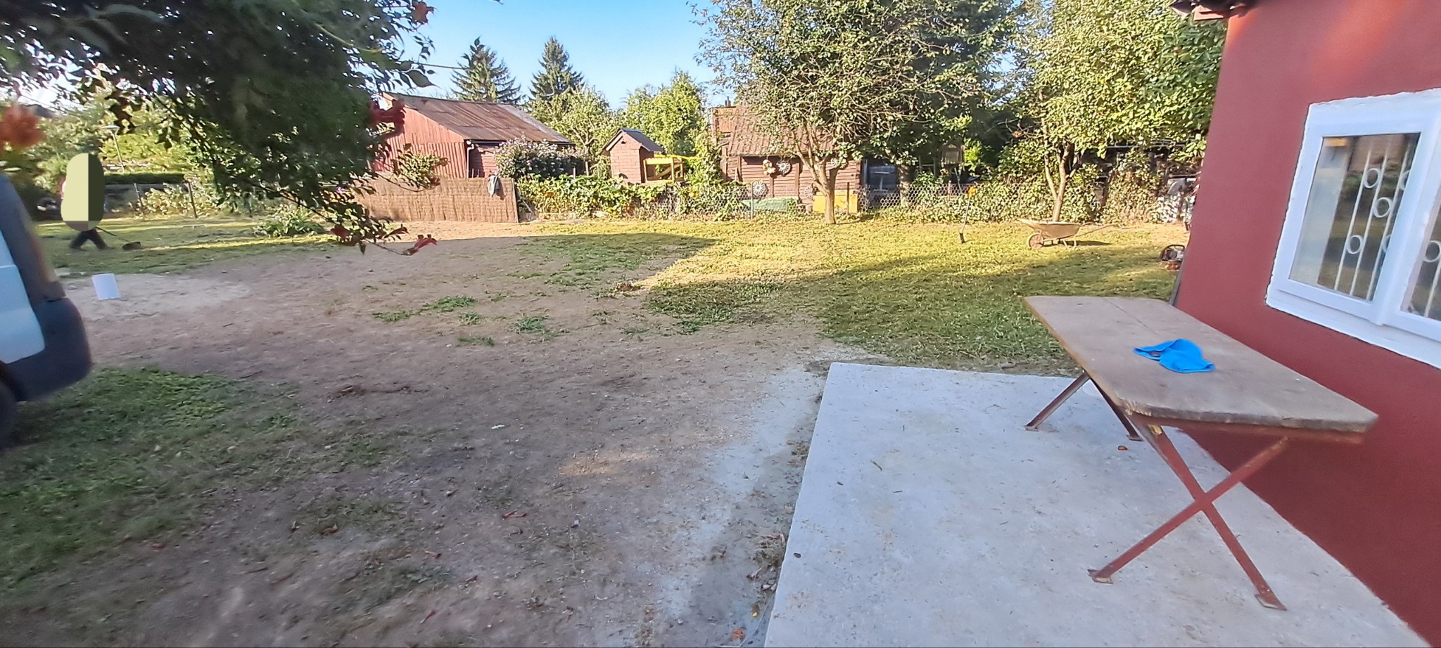 Rozległy teren posesji z drewnianymi budynkami gospodarczymi w tle, widok z bliska na stół roboczy z niebieską szmatką, sugerujący prace porządkowe lub przygotowawcze.