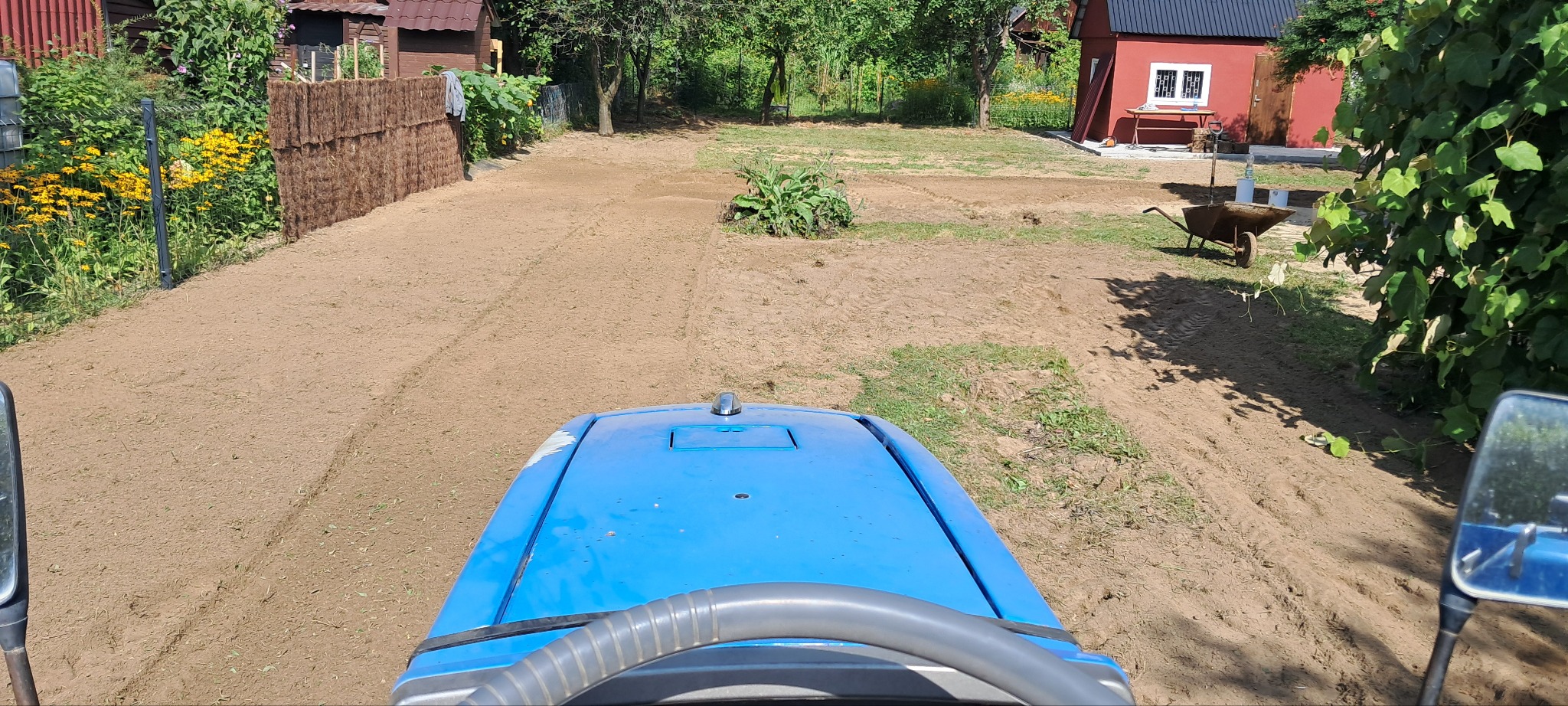 Widok z kabiny traktora na świeżo wyrównany teren działki z widocznymi śladami opon, przygotowany pod przyszłe nasadzenia. W tle zabudowania gospodarcze i zieleń.