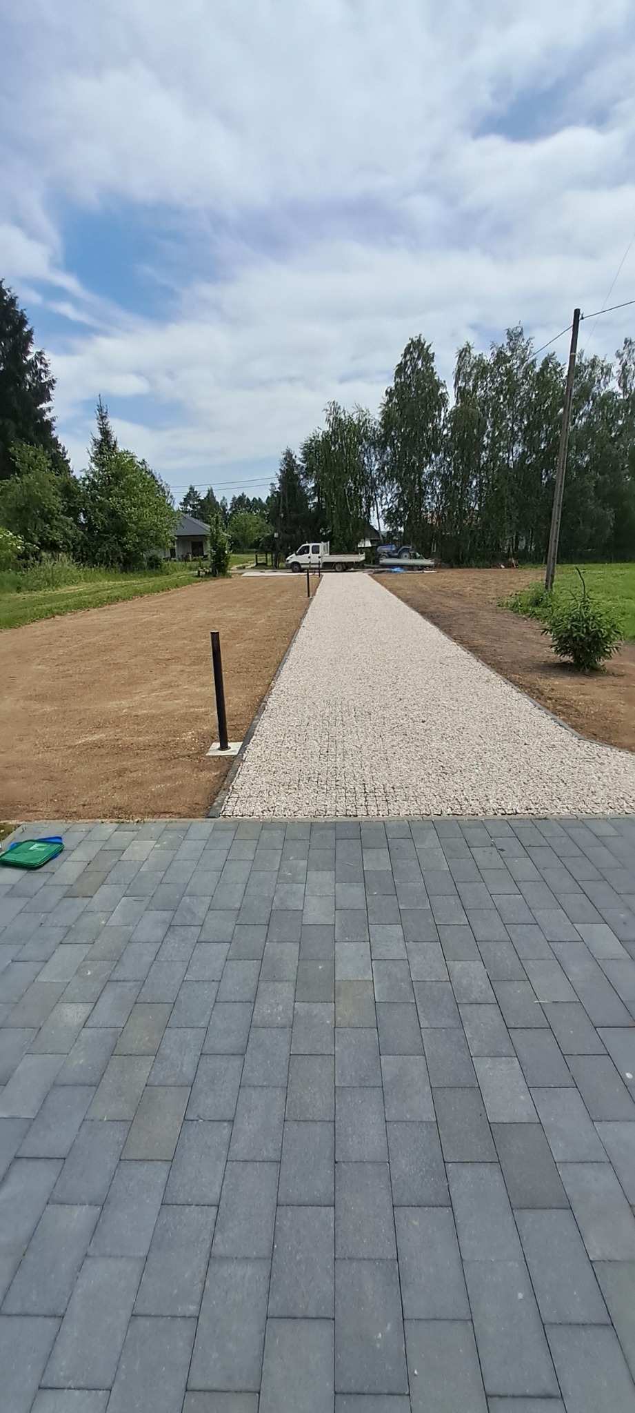 Ukończona ścieżka z jasnego żwiru prowadząca przez trawnik, zakończona szarą kostką brukową. W tle widoczny samochód dostawczy i drzewa.