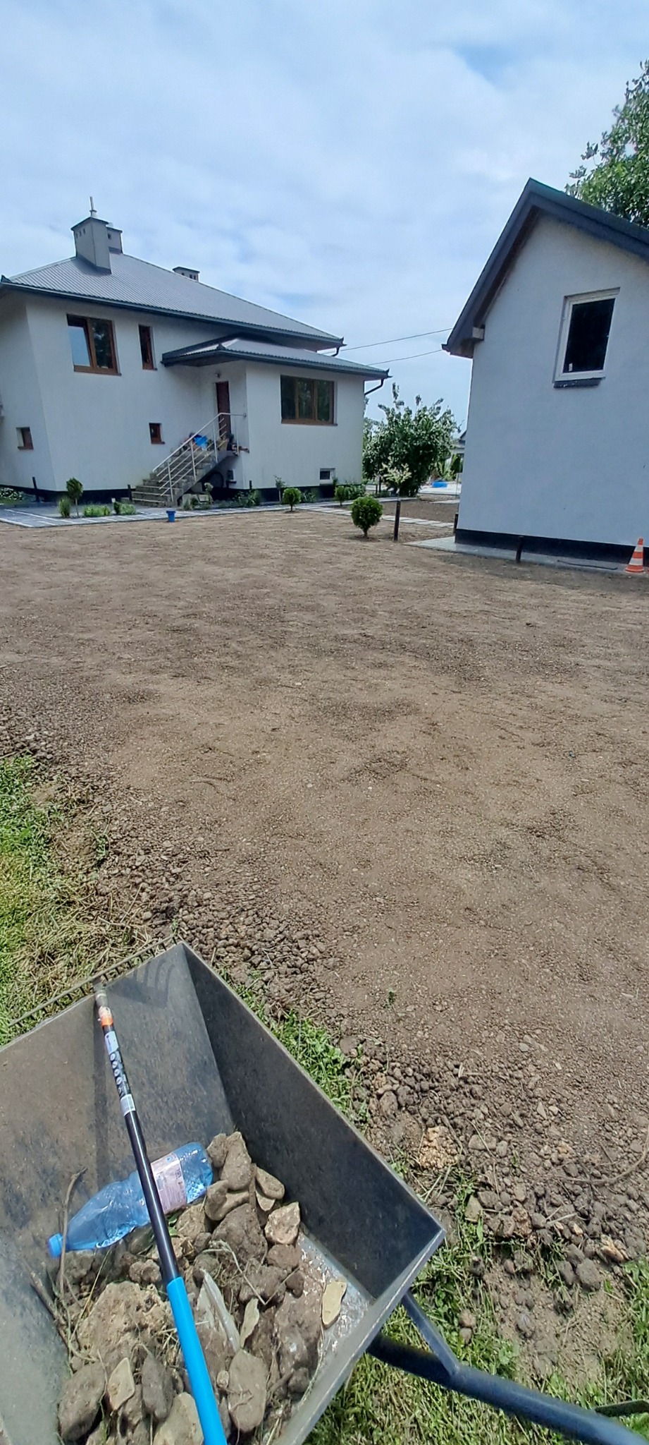 Wyrównany teren działki z widocznymi dwoma budynkami w tle. W pierwszym planie taczka z kamieniami i narzędziem. Widok na posesję po niwelacji.