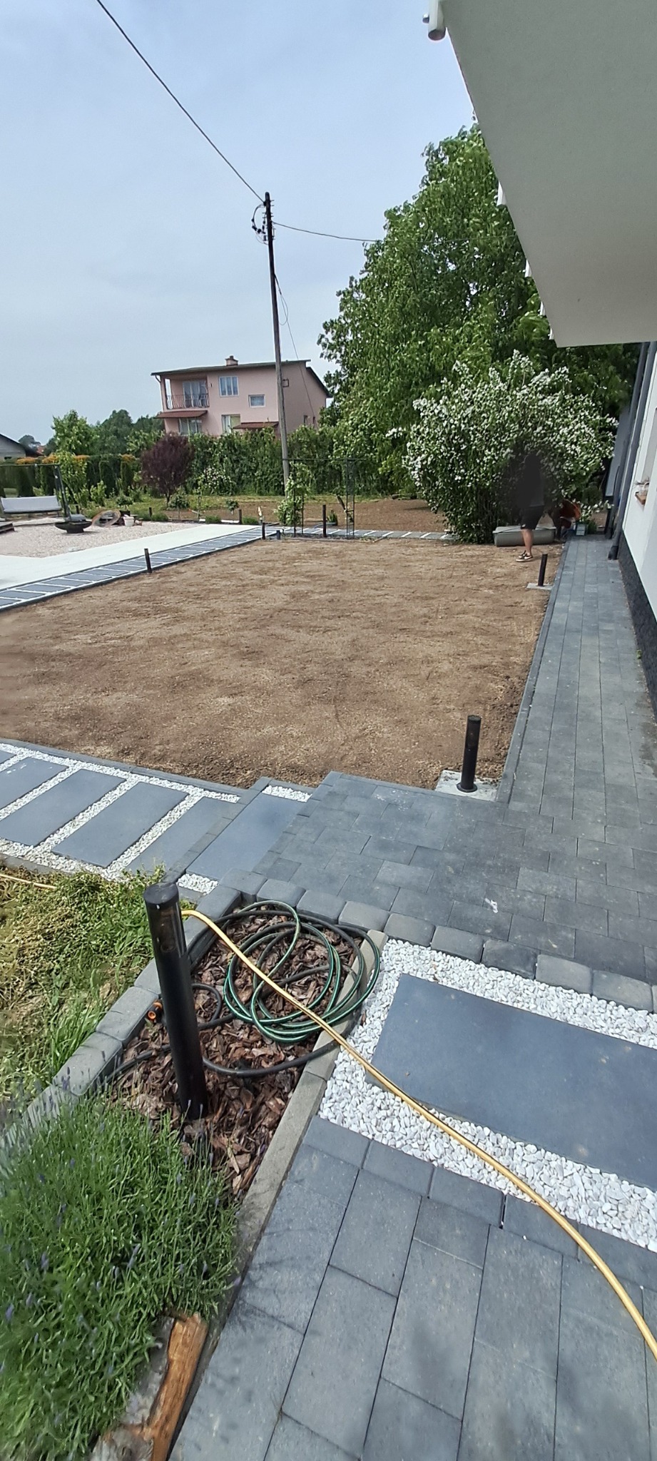 Przygotowany teren pod trawnik z nawodnieniem, widok z boku domu. Kostka brukowa, rabaty z korą i roślinami. W tle dom i zieleń.