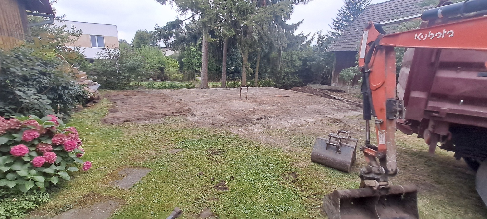 Przygotowany teren pod budowę z widocznym sprzętem budowlanym Kubota, otoczony zielenią i drzewami. W tle budynki mieszkalne.
