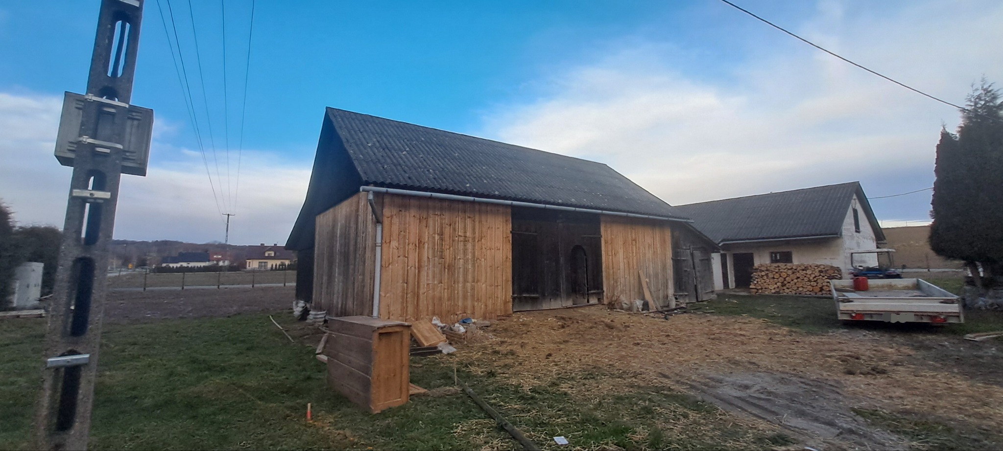 Odnowiona drewniana stodoła z szarym dachem, widoczna na tle wiejskiego krajobrazu. Częściowo wymienione deski elewacyjne. Na pierwszym planie skrzynia i ziemia.