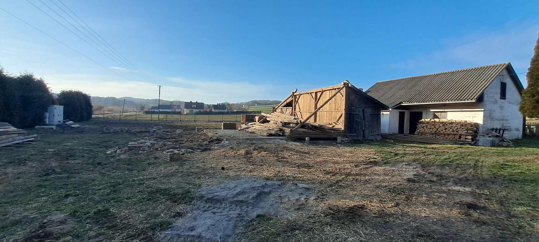 Częściowo rozebrany drewniany budynek gospodarczy na tle błękitnego nieba i zabudowań wiejskich. Widoczne sterty drewna i przygotowany teren.