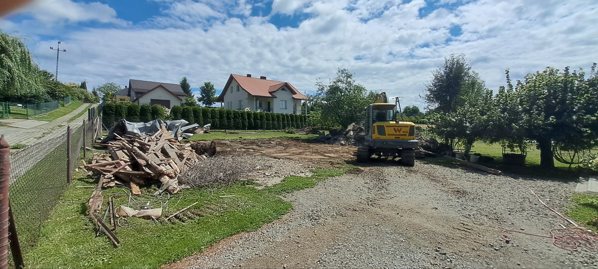 Żółta koparka Wacker Neuson na terenie budowy, z stertą gruzu i wykopem w tle, pod błękitnym niebem z chmurami. Widoczne domy i zieleń.