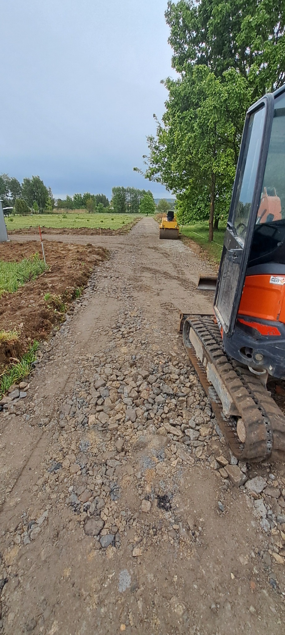 Utwardzanie drogi gruntowej z użyciem walca i minikoparki. Widoczne świeżo rozplantowane kamienie i ziemia. Prace w toku, sprzęt budowlany w użyciu.