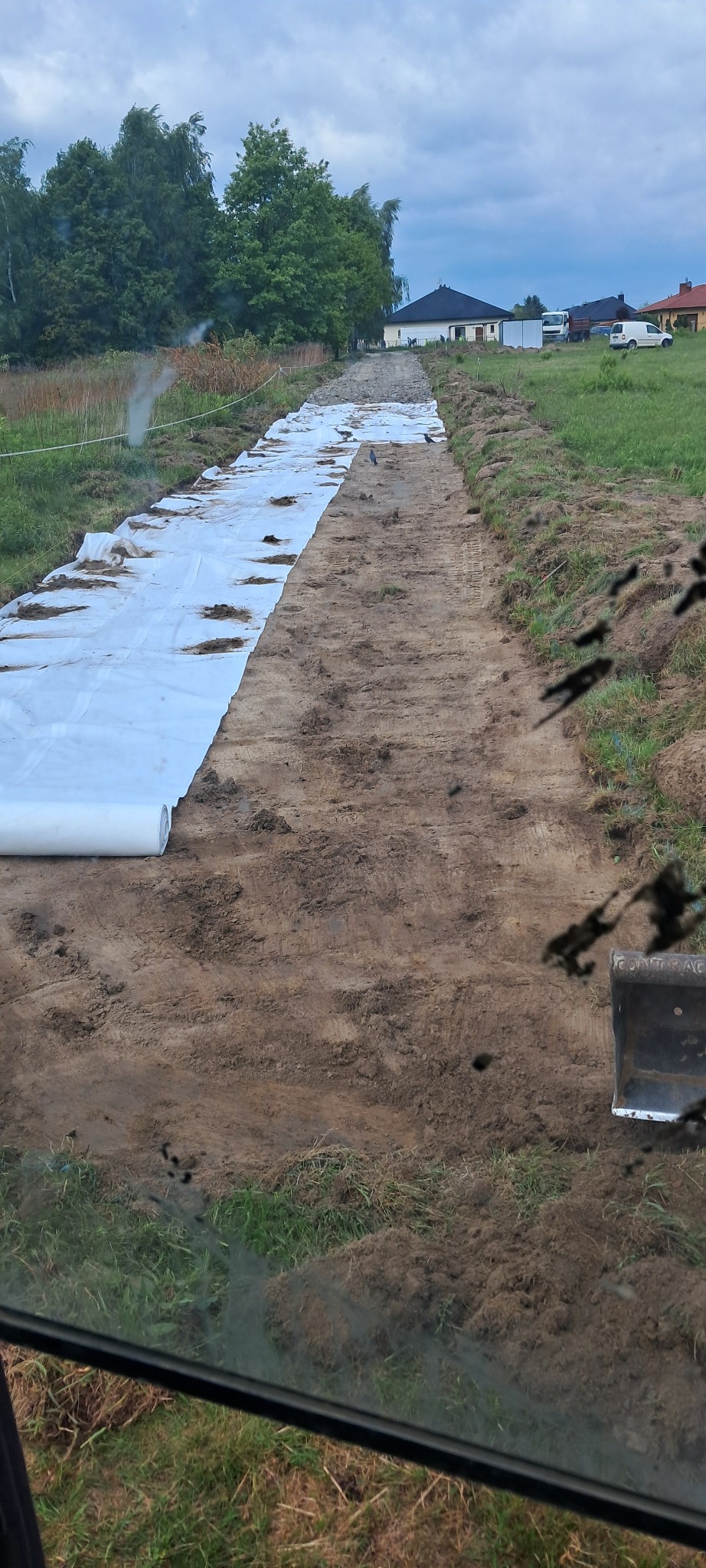 Przygotowanie podłoża pod drogę: rozłożona biała geowłóknina na wyrównanym terenie, widok z kabiny maszyny budowlanej, w tle zabudowania.