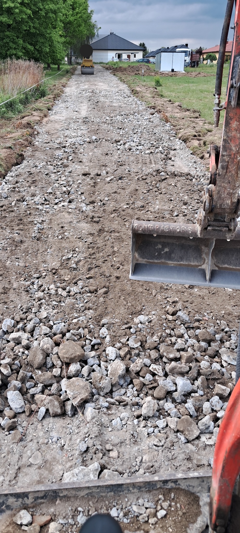 Utwardzanie drogi kruszywem z użyciem koparki i walca drogowego. Widok z perspektywy operatora koparki na plac budowy drogi gruntowej.