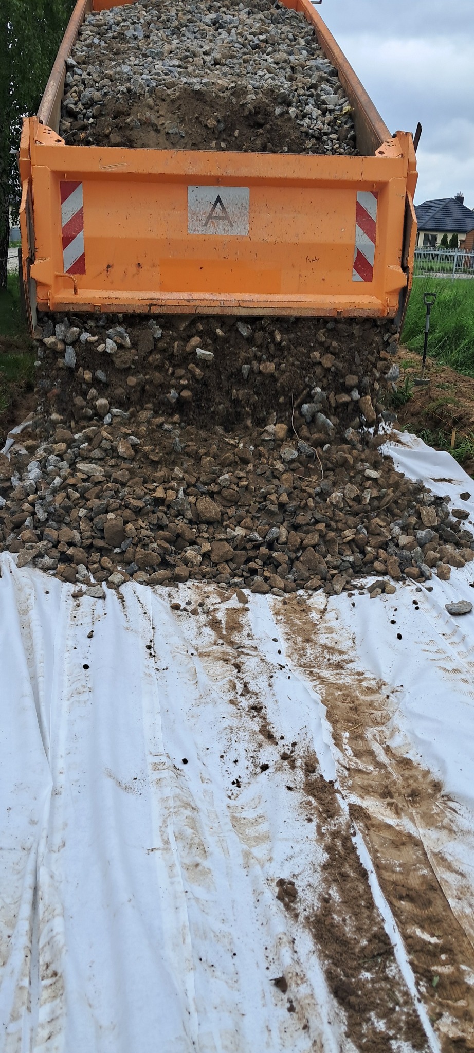 Pomarańczowa wywrotka wysypuje gruz na białą geowłókninę. Widoczne domy w tle i fragment drzewa. Utwardzanie terenu pod budowę.