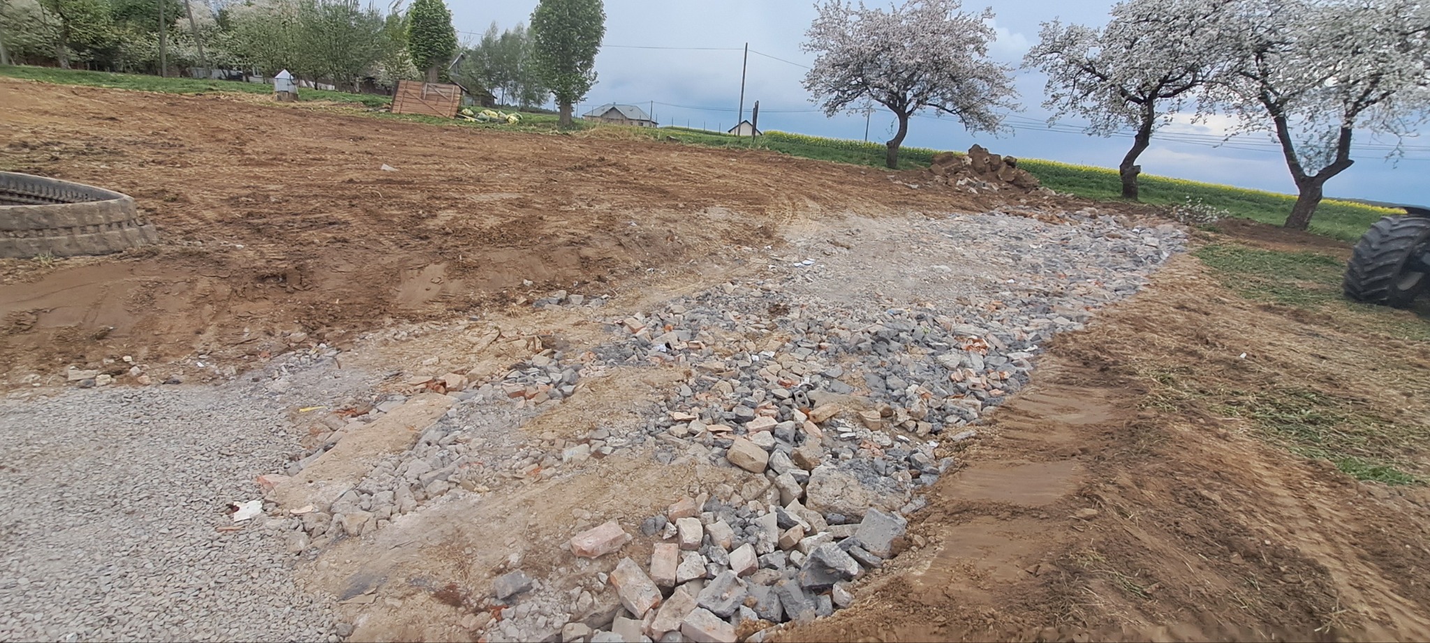 Przygotowanie terenu pod budowę drogi: wyrównany teren z gruzem i kamieniami, w tle drzewa owocowe i pole rzepaku. Widoczne ślady opon traktora.