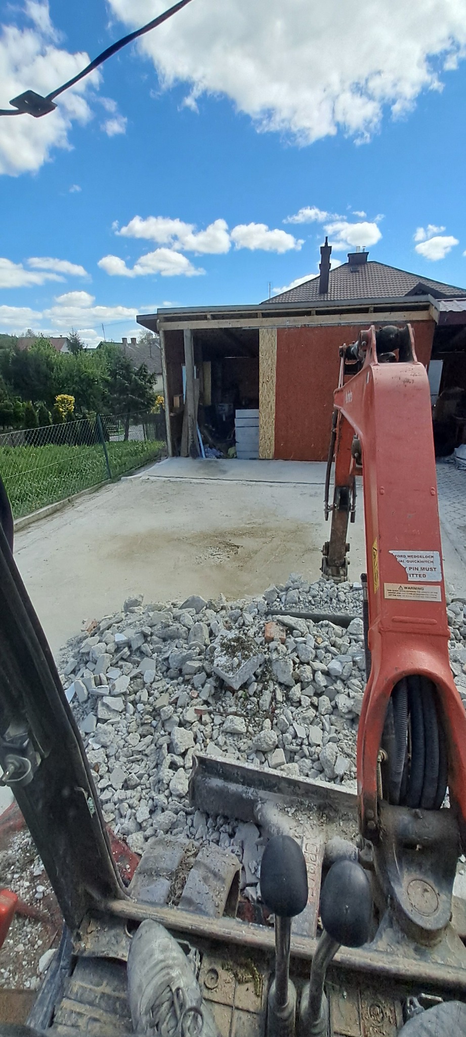 Widok z kabiny minikoparki na gruzowisko po rozbiórce, z budynkiem w tle i błękitnym niebem. Fragment maszyny widoczny na pierwszym planie.