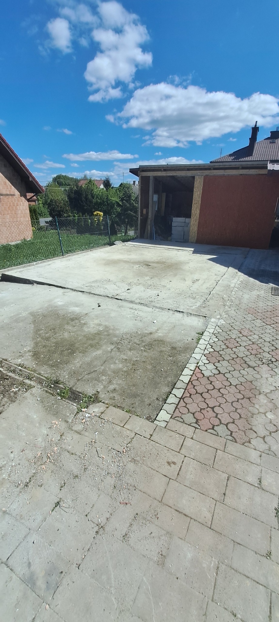 Betonowy plac przed garażem z kostką brukową, ogrodzeniem i budynkami w tle, w słoneczny dzień. Widoczne ślady po pracach budowlanych.