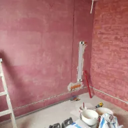 Prace wykończeniowe wewnątrz pomieszczenia: ściany pokryte różowym tynkiem, widoczne puszki elektryczne, przewody, drabina malarska, narzędzia i worki z zaprawą Knauf MP 75 L.