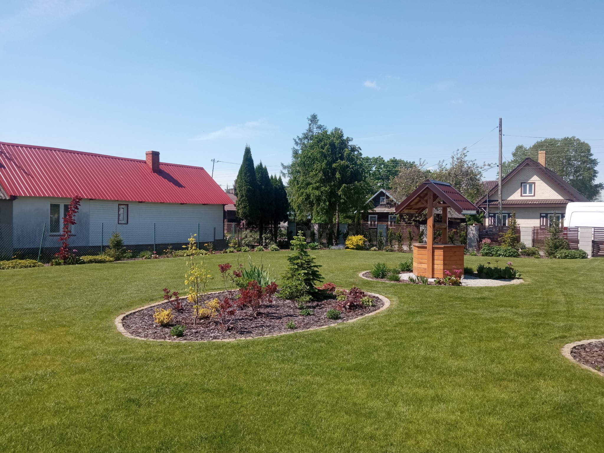 Utrzymany ogród z równo przystrzyżonym trawnikiem, rabatami z korą i roślinami oraz drewnianą studnią pośród zabudowy jednorodzinnej w słoneczny dzień.