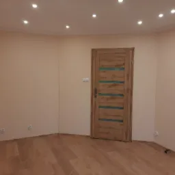 Wnętrze pokoju po remoncie: ściany pomalowane na jasny beż, drewnopodobna podłoga, zamontowane drzwi z drewnianą okleiną i poziomymi, przezroczystymi paskami, oświetlenie sufitowe punktowe.